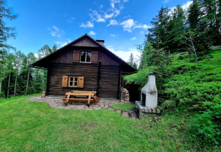 Holzchalet Schneiderhütte in Österreich, umgeben von grüner Natur, Bäumen und blauem Himmel.