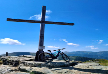Cykel ved stort trækrucifiks på Schneiderhütte i Østrig, med bjerge og blå himmel i baggrunden.
