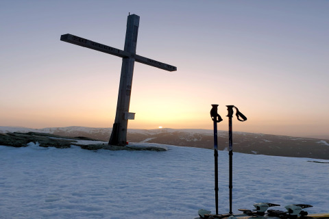 Sonnenuntergang bei der Schneiderhütte in Österreich mit Holzkreuz, Skistöcken und verschneiten Bergen.