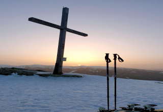 Sonnenuntergang bei der Schneiderhütte in Österreich mit Holzkreuz, Skistöcken und verschneiten Bergen.