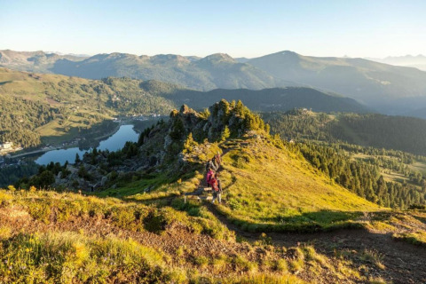 Aussicht von der Schneiderhütte in Österreich mit Wanderern, Bergen, See und sonnigem Himmel im Hintergrund.