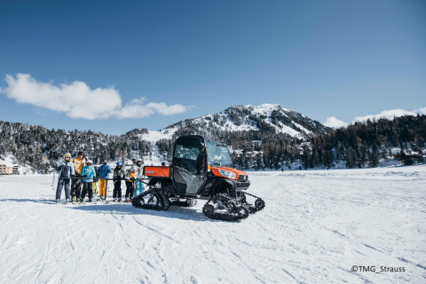 Group of skiers and a tracked vehicle in the snow at Alpenpark Turrach 21 in Austria, mountains behind.