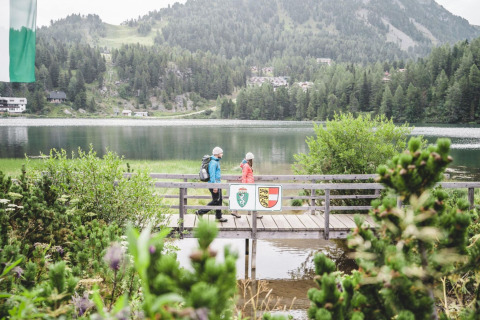 Two hikers cross a wooden bridge by the lake at Alpenpark Turrach 21 in Austria, surrounded by mountains.