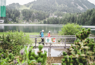 Two hikers cross a wooden bridge by the lake at Alpenpark Turrach 21 in Austria, surrounded by mountains.