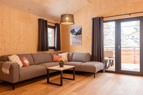 Moderne woonkamer in een boomhut met grijze bank, houten wanden, grote ramen en uitzicht op de natuur.