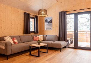 Moderne woonkamer in een boomhut met grijze bank, houten wanden, grote ramen en uitzicht op de natuur.