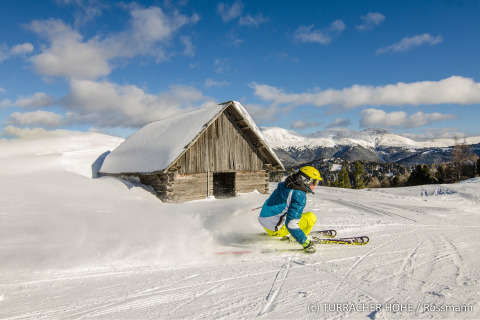 Skiër glijdt langs een met sneeuw bedekt boomhut-chalet in Turracher Höhe, Oostenrijk, met bergen erachter.