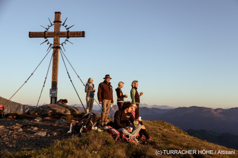 Hiking group enjoys sunset by a large cross on a mountain peak at Turracher Höhe in Austria.