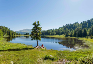 Peaceful lake at Alpenpark Turrach 21 in Austria, surrounded by green hills and pine forests, sunny sky.