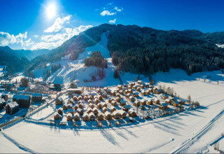 Verschneite Chaletsiedlung Kreischberg Chalets 21 vor bewaldeten Bergen, Sonnenlicht und blauem Himmel.