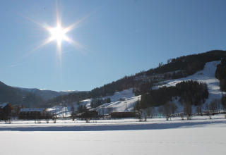 Zonnig winterlandschap met besneeuwde heuvels en bomen, gezien vanuit een lodge met huisjes.