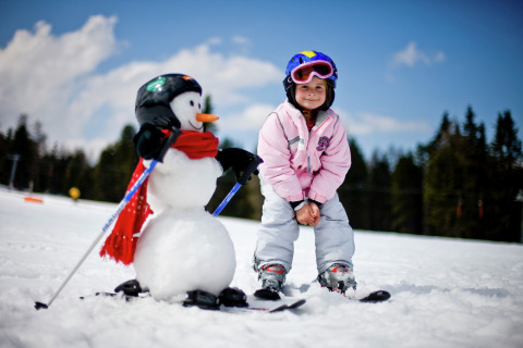 Ein Kind in Skibekleidung und Helm steht neben einem Schneemann mit Skiern und Helm im Schnee.