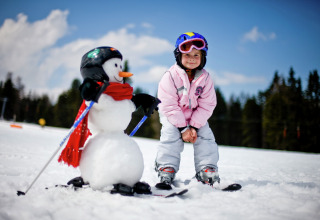 Een kind in skikleding en helm staat op ski’s naast een sneeuwpop met ski’s en helm op een besneeuwde helling.