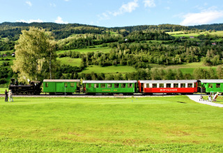 Een ouderwetse stoomtrein rijdt langs een glamping-accommodatie met groene heuvels op de achtergrond.