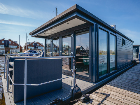 Modernes Hausboot Havenlodge Plus mit großer Fensterfront und Terrasse, vertäut am Steg bei Sonnenschein.