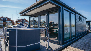 Moderne husbåd Havenlodge Plus med store vinduer og terrasse fortøjet ved en bådebro under en klar himmel.
