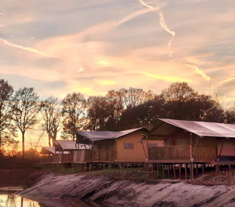 Safari tent for 6 people with sanitary facilities by the lake, photographed at sunset with trees in the background.