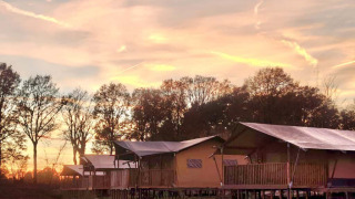 Safari tent for 6 people with sanitary facilities by the lake, photographed at sunset with trees in the background.