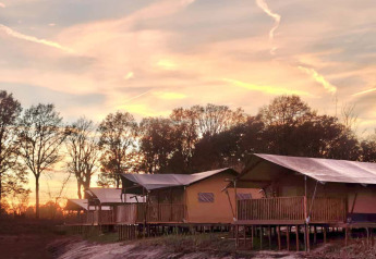 Tente safari pour 6 personnes avec sanitaires au bord du lac, prise au coucher du soleil avec des arbres en arrière-plan.