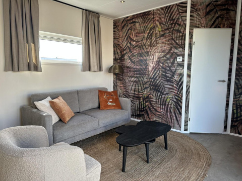 Moderne leefruimte in de Zonneweelde Lodge Deluxe bij Camping Zonneweelde, met grijze zetel en kunstmuur.