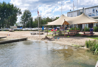 Beach scene with glamping tents, picnic tables, and boats by the water on a partly cloudy day.