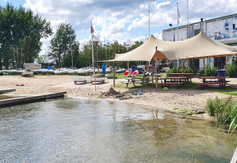 Scène de plage avec zone de glamping, grands tentes, tables de pique-nique et bateaux près de l’eau.