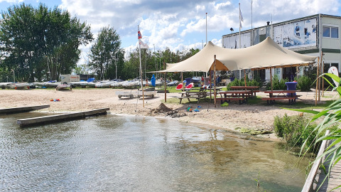 Escena de playa con zona de glamping, grandes carpas, mesas de picnic y barcos junto al agua.