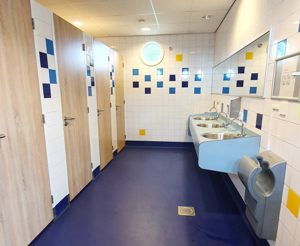 Baño público moderno en Marina Parcs Almere, con suelo azul, lavabos de acero y cabinas privadas.