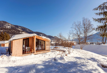 Ein charmantes Holzchalet steht im Schnee, umgeben von Bergen und Bäumen unter klarem, blauem Himmel.