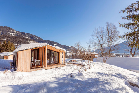 Et charmerende chalet i træ står dækket af sne, omgivet af bjerge og træer under en klar blå himmel.