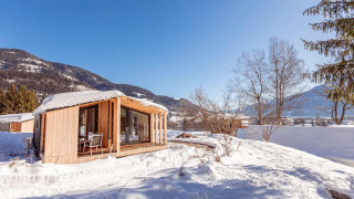 Een gezellig houten chalet bedekt met sneeuw, omgeven door bergen en bomen onder een stralend blauwe lucht.