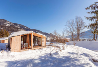 Et charmerende chalet i træ står dækket af sne, omgivet af bjerge og træer under en klar blå himmel.