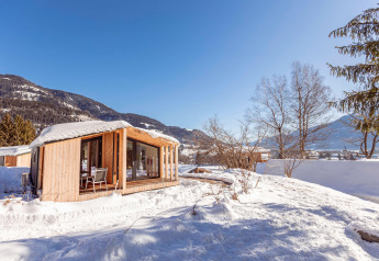 Un accogliente chalet in legno coperto di neve, circondato da montagne e alberi sotto un cielo blu brillante.