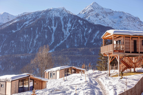 Sfeervolle houten chalets in de sneeuw met indrukwekkende bergen op de achtergrond onder een blauwe lucht.
