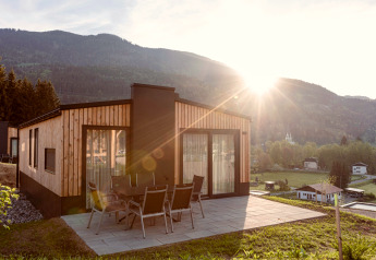Modernes Holzchalet mit Terrasse und Gartenmöbeln, Berglandschaft und Sonnenschein im Hintergrund.