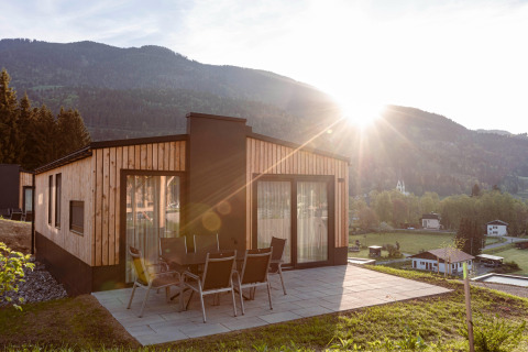 Houten chalet met terras en tuinset, berglandschap en zonsondergang op de achtergrond zichtbaar.