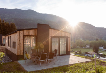 Chalet in legno con patio e tavolo da esterno, paesaggio montano e sole che sorge sullo sfondo.