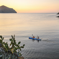 Twee mensen kajakken bij zonsondergang op rustig water, omgeven door bomen in een glampingpark.