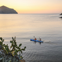 Twee mensen kajakken bij zonsondergang op rustig water, omgeven door bomen in een glampingpark.