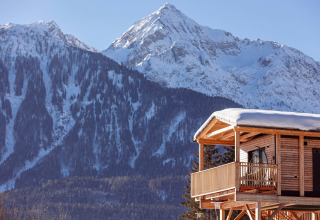Houten chalet met besneeuwd dak en balkon in Oostenrijk, met besneeuwde bergen op de achtergrond.