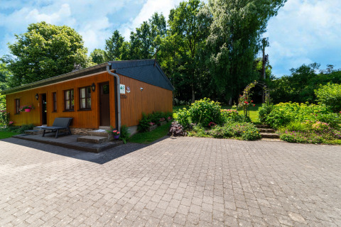 Houten lodge 'Les Bouleaux' in België, omgeven door tuin, bloemen en groene bomen onder blauwe lucht.
