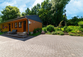 Houten lodge 'Les Bouleaux' in België, omgeven door tuin, bloemen en groene bomen onder blauwe lucht.