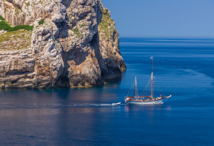 A sailing boat cruises by rocky cliffs near Alghero in Sardinia, Italy, on a calm, clear blue sea day.