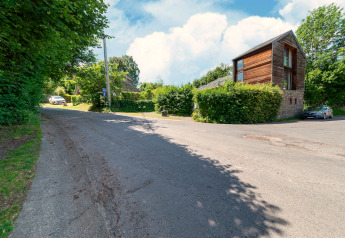 Photo of 'Les Bouleaux' lodge in Belgium, modern wood and stone building beside a quiet road and greenery.