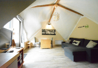 Stue i chalet 'Mise Au Vert' i Belgien med skrå loft, stor sofa, tv og spiseområde under træbjælker.