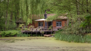 A cozy wooden cabin named The Wijerschans by a pond, surrounded by lush green forest and vegetation.