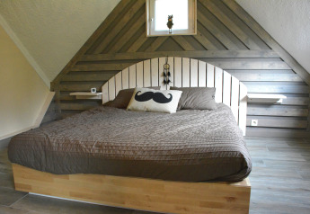Soveværelse med træpaneler, stor seng, brun dyne og en pude med overskæg i chalet Mise Au Vert, Belgien.