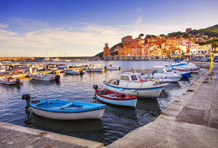 Barcos atracados en el puerto de Lacona, con casas coloridas y colinas de fondo en la Toscana, Italia.