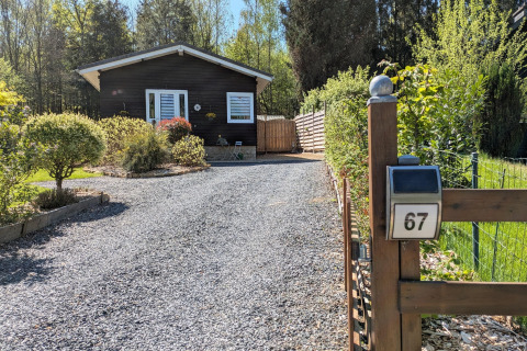 Oprit van grind naar lodge Chatillon 67 in België, houten huisje met veel groen en voorjaarszon.