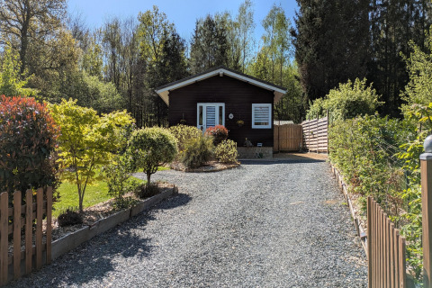Ingang naar lodge 'Chatillon 67' in België met grindpad, groene planten en houten huisje op de achtergrond.